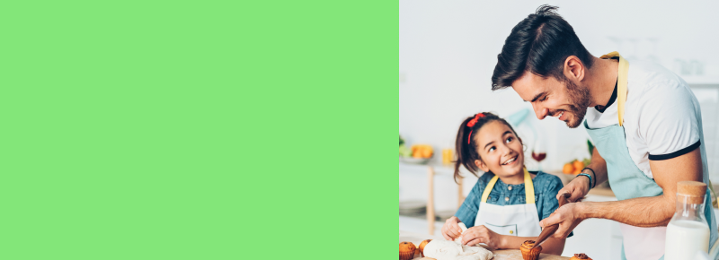 Father and daughter baking together in a bright kitchen, smiling and enjoying the activity; the girl is shaping dough while the father decorates muffins, with cookies, cookie cutters, and ingredients visible on the counter.