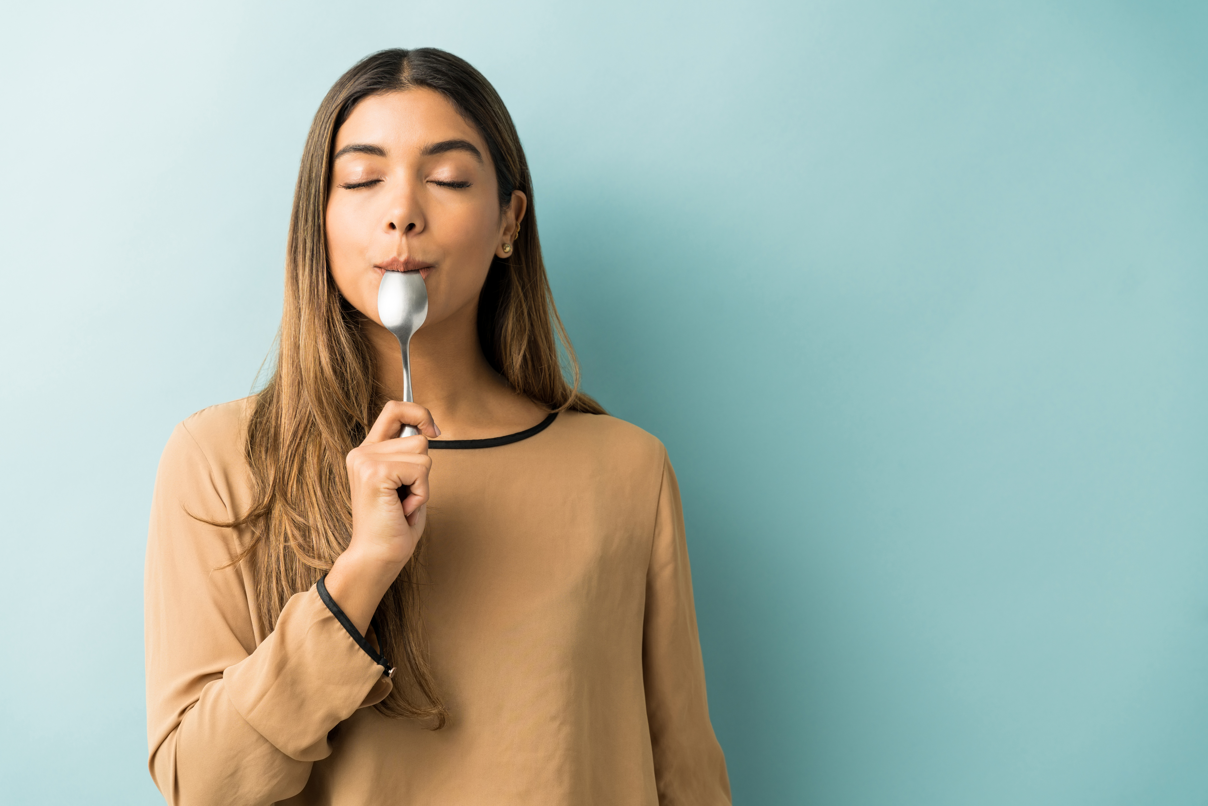 Women with spoon in mouth, eyes closed, looking happy and satisfied against a blue wall.